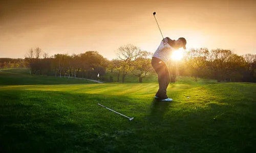 a lone golfer chips onto the green , the low sun is coming from behind him and flaring to camera.